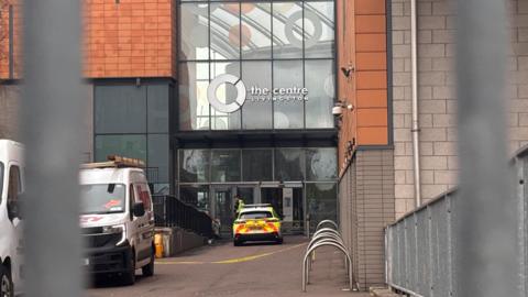 An entrance to The Centre Livingston at Almondvale Shopping Centre, showing a police vehicle and a white van parked near the glass-fronted doorway.