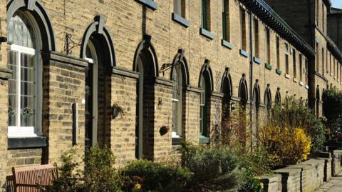 A row of terraced houses, built of Yorkshire stone, all with neat front gardens and hedges, uniform doors and windows and benches lining the street.