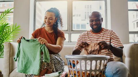 A woman and man sit on a sofa folding clothes into a washing basket.