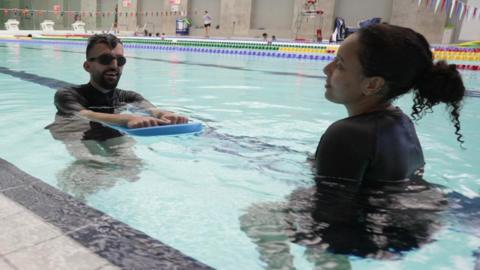 A man is holding a float in a swimming pool. He is standing in front of a woman who is teaching him to swim.