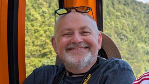 Neil Bradley with glasses atop his head smiling inside a cable car going past a hill covered in trees