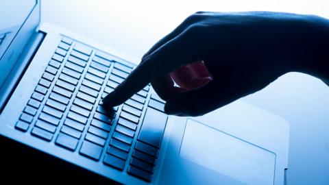 The photo shows a hand with a finger on a key on a laptop. There is a blue tint to the image and the hand is a silhouette. 