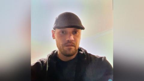 Atis Bleive is in the middle of the image. He is wearing a black hat, T-shirt and coat, and is taking a selfie of himself. 