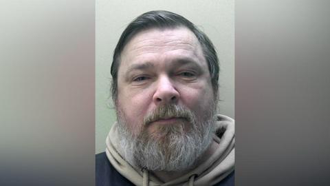A headshot of a man with a grey beard wearing a beige hoodie underneath a black coat.