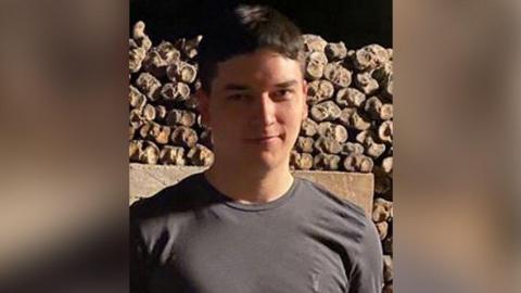 Tom Dingle with short brown hair wearing a grey t-shirt stands in front of a stone wall in the open air. It is evening. He is smiling.