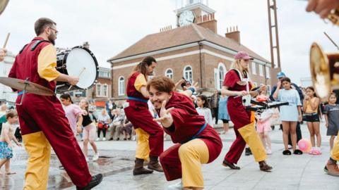A performing group dancing and playing musical instruments on the street in front of a crowd. One of the performers is smiling while holding a hand out to the camera.