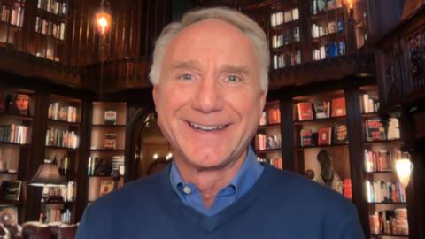 Dan Brown smiles. He has short grey hair and wears a blue shirt under a blue jumper. He is in a room full of bookshelves