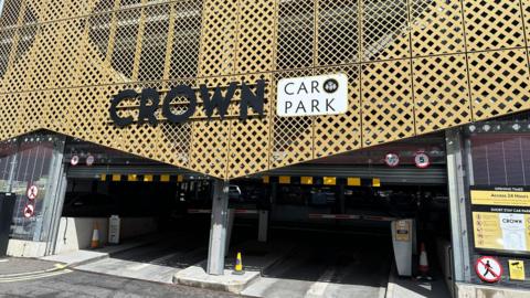 The outside of a multi-storey car park, a sign reads Crown Car Park. A yellow facade is on the outside walls of the building. Two barriers block two roads into the car park.