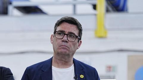 Headshot of Burnham who looks up and to the right. He has a serious expression. The background is blurry.