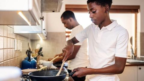 Stock photo showing a teenager using a fying pan while cooking in the kitchen with his father in the background