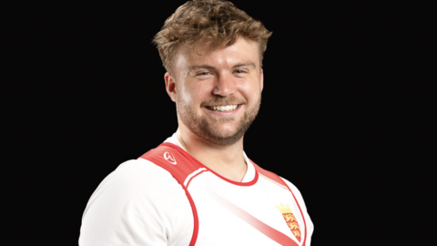 Olly Dodds is smiling at the camera and is wearing a red and white weightlifting vest over a white t-shirt.