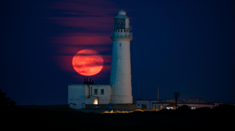 A red-coloured moon to the left of a white lighthouse. It is night time and the sky is a dark blue.