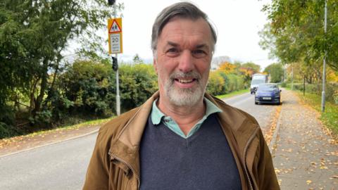 Rupert Pearce-Gould smiling into camera next to a road in Harston with a school 20mph limit in the background. He has grey hair swept to the side, a goatee beard, is wearing a brown jacket, navy jumper and turquoise shirt.