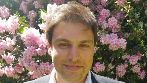 Adam Dodd, a man with short brown hair, is pictured in a posed headshot in front of a bush with pink flowers.