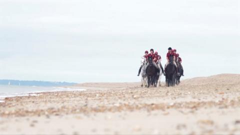 A group of black and white horses are gathered together running along the beach, their riders wear burgundy t shirts and black helmets