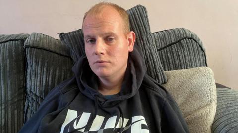 Jonathan Johnson, a man with short blond hair wearing a black Nike hoody, sits on a sofa 