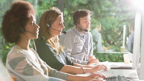 A man and two women in front of a computer typing - the man is wearing a shirt, one woman wearing a green jumper, another woman wearing a pink and green top. They are all wearing headsets. 