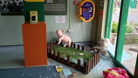 The image shows the scene of a break-in. It is in the foyer of a farm where a large glass door has been smashed and glass is all over the floor. A fake farm scene with a plastic pig and a goat is all thrown around and there is glass all over the artificial grass. 