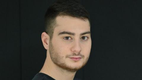 A photograph of a young man with dark hair, well lit in a dark room