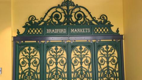 Decorative iron panels with scrollwork and the words Bradford Markets in gold against a yellow wall