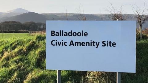 A white sign in front of a grass mound with hills in the background