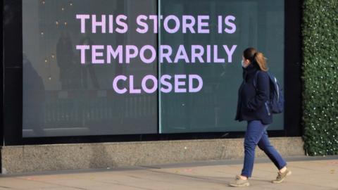 Woman in mask walks past shop sign saying 'This store is closed'