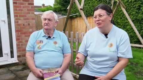 Charles and Tina wearing matching blue T-shirts and sitting in a garden with a lawn and a swing behind them