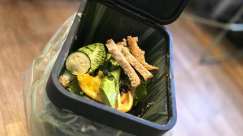 A food waste bin. The black caddy has the lid open showing food waste, including bread, lettuce and other vegetables in a clear bin bag inside it.