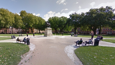 Queen Square, Bristol. People are sat on benches in a semi circle, with trees and grass around them