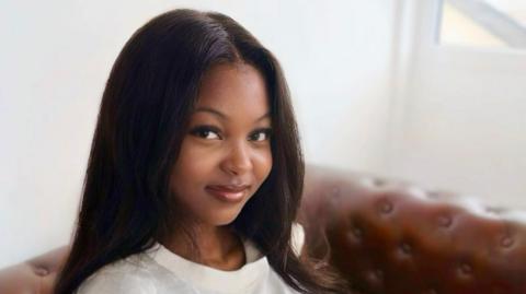 A headshot of a woman who is sat on a sofa looking at the camera. She is smiling and has long brown hair. The background is blurred.