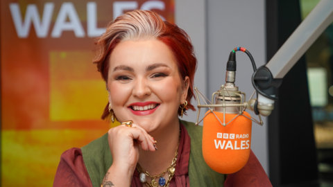 A woman with short died red hair, wearing a red shirt and green waistcoat and smiling at the camera. She has her chin leaning on one hand, and is wearing gold earrings, rings and necklace. In front of her is an orange microphone with BBC Radio Wales written on it.