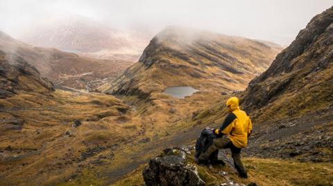 A person in waterproof clothing stands looking out onto a wet and cloudy landscape in the Scottish highlands, with mountains and rivers in their view