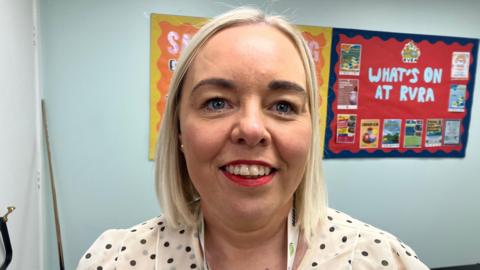 A woman with shoulder length blonde hair, wearing a white top with black dots on it. There is a red poster on the blue wall behind her, reading 'what's on at RVRA'. 