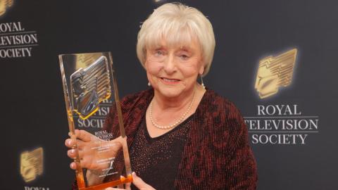 Ann Ming holds a large glass trophy in front of a black banner with the Royal Television Society written on it. She is elegantly dressed in a dark red and black jacket, with short white hair.