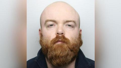 Benjamin Pinchin with a bald head and thick ginger beard. He is wearing a black top and has a stern expression as he stands in front of a grey background.