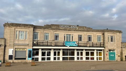 Old seaside building on the seafront. It is a classic opening to an old lido on a road. There is a big sign at the top that says "Tropicana" with a cloudy, moody sky.