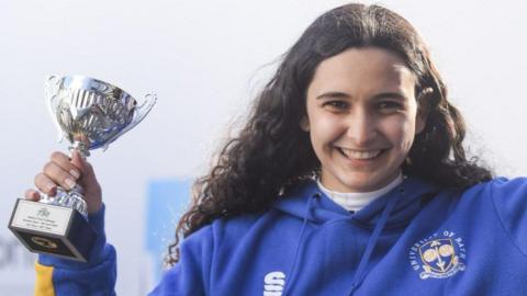 Stephanie wearing a blue hoodie with long curly brown hair smiling at the camera. She is holding an award.