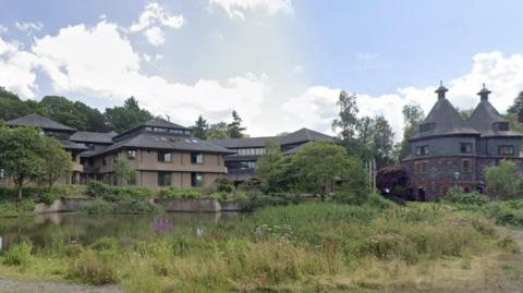 Powys County Council's headquarters in Llandrindod Wells. It is a two-storey brown brick building with a dark-coloured tiled roof. There are several buildings of the same style surrounding it and they sit next to a pond with trees dotted around.