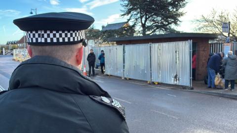 Metal fencing outside the Sheringham bus shelter where campaigners have been gathered. In the foreground of the picture is a police officer. The picture has been taken over the officer's shoulder who is wearing police uniform and a hat.