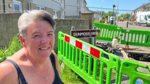 A woman with short grey hair stands in front of a green barrier