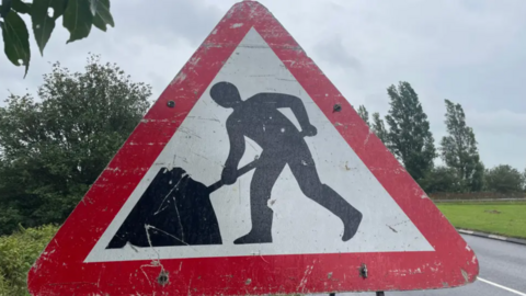 A large triangle roadworks sign with greenery behind it.