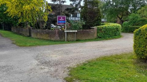 An entry to a gravel road, with a sign saying Tudor Place against a low brick wall. There is grass on either side of the gravel and a black and white house in the background through trees