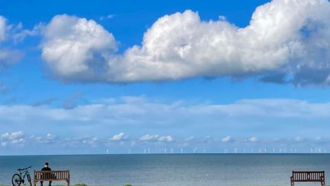 A cyclist sits on a bench looking out to sea under a blue sky with white clouds, with wind turbines on the horizon