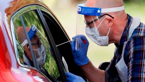A man holding a swab takes a a test from someone in a car.