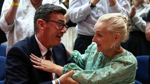 Andy Burnham hugging his wife, Marie-France van Heel. Burnham is wearing a dark suit with a shirt and tie and glasses. Van Heel is wearing a green dress. She has blond hair and hooped earrings.
