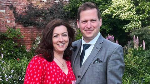 Ben and Sarah Howard standing in a garden. Sarah is wearing a red dress with an all-over spot pattern and holding a drink. Ben is wearing a grey suit and dark tie