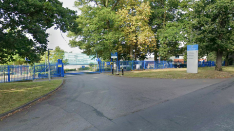 The entrance to the school has blue gates and trees surrounding it with a wide entrance form the road.
