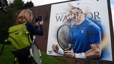 Woman carrying a green backpack and a tennis racket uses her phone to take a photograph of a large artwork of Andy Murray. It is a picture of Murray in a blue t-shire and white cap. It says Andy Murray. Warrior. Legend. 