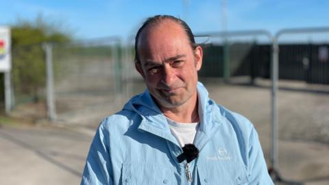 A man in a blue coat is standing in front of security fencing