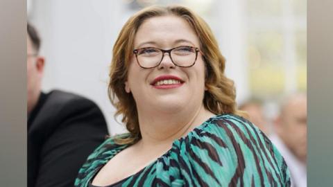 A woman in a black and green striped dress smiles while looking into the distance. She is wearing glasses.